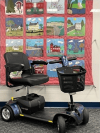 A mobility scooter is parked indoors in front of a colorful quilt decorated with scenes of buildings, trees, and landscapes, each framed by pink borders.