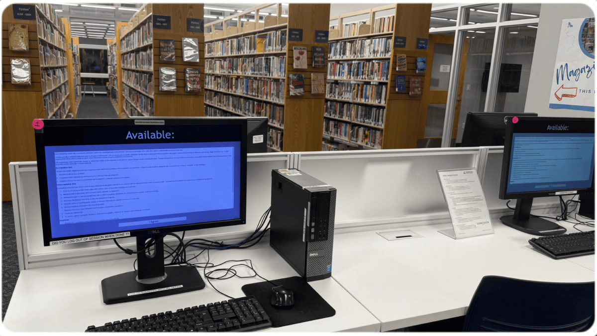 A library computer workstation with a desktop computer and monitor displaying “Available,” a keyboard, and mouse. Bookshelves filled with books are visible in the background. Another computer is at a nearby desk.