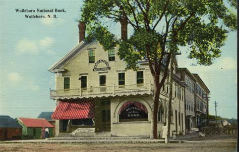 A vintage postcard of the Wolfeboro National Bank in Wolfeboro, New Hampshire shows a large, three-story building with red and white striped awnings, trees nearby, and a clear sky.